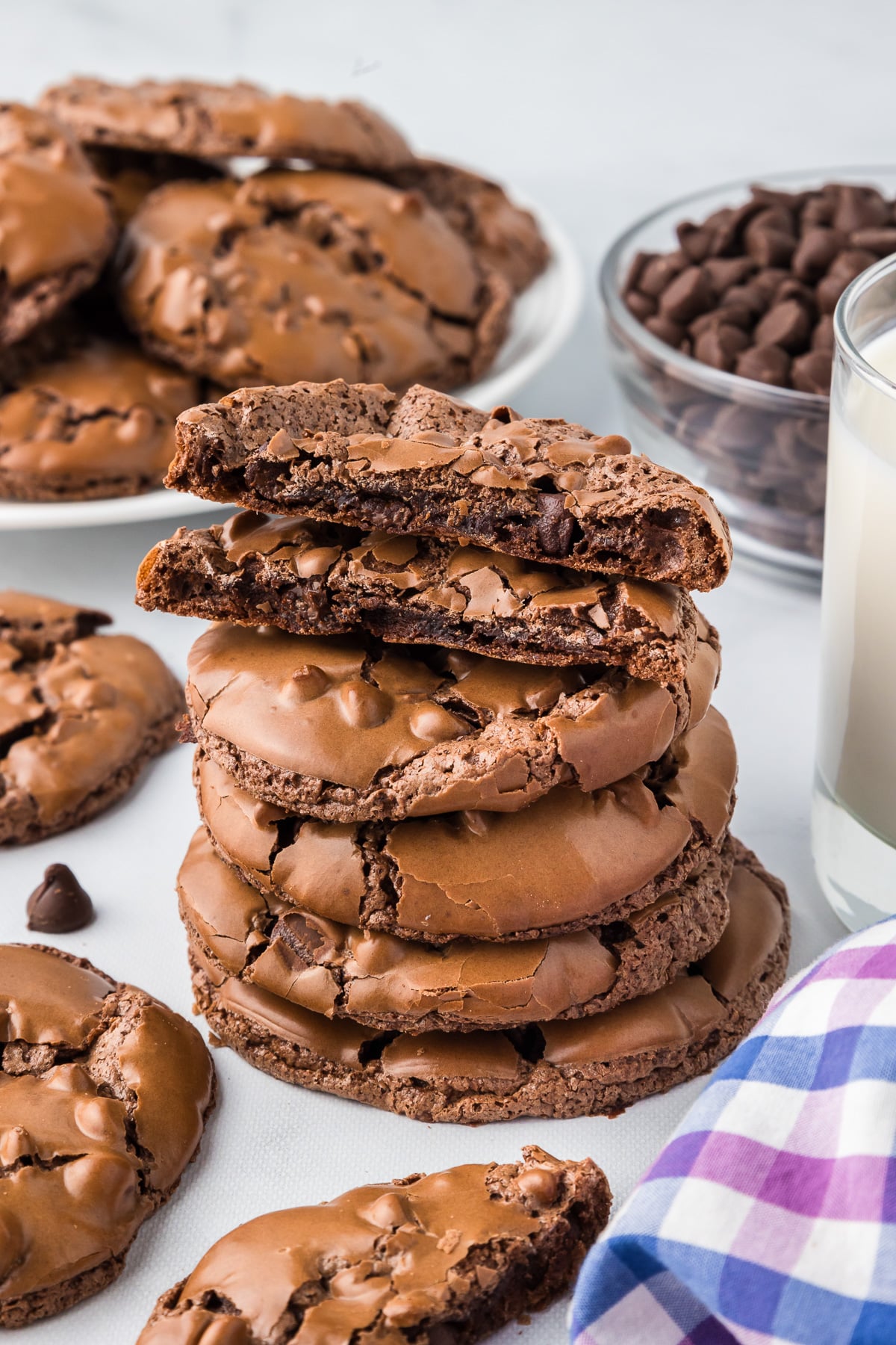 A stack of flourless chocolate brownie cookies with the top cookie cut so you can see the inside, with a glass of milk and more cookies on a plate in the background.
