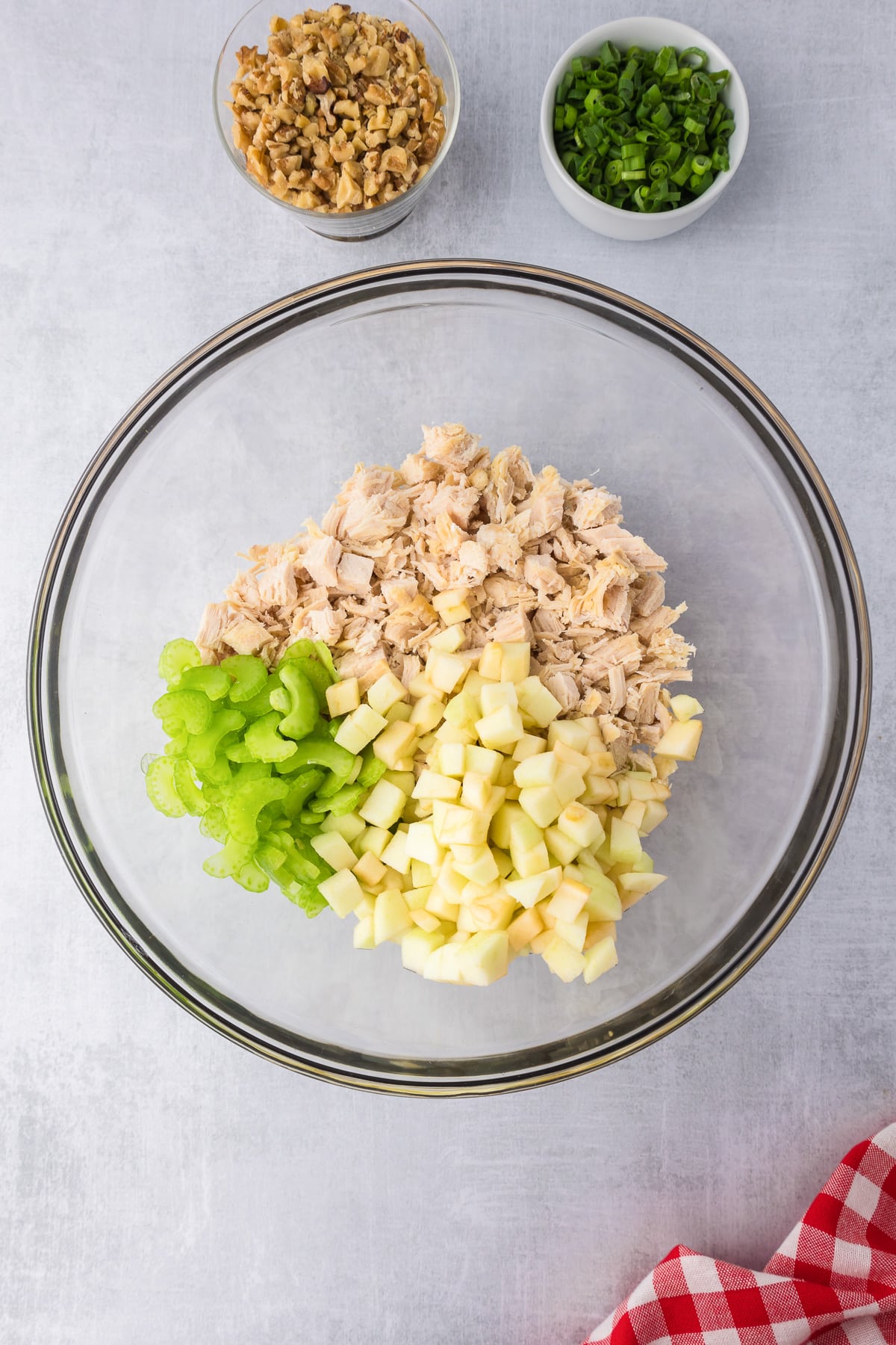 A glass bowl filled with chopped chicken, onions and celery with walnuts and green onions in bowls nearby.