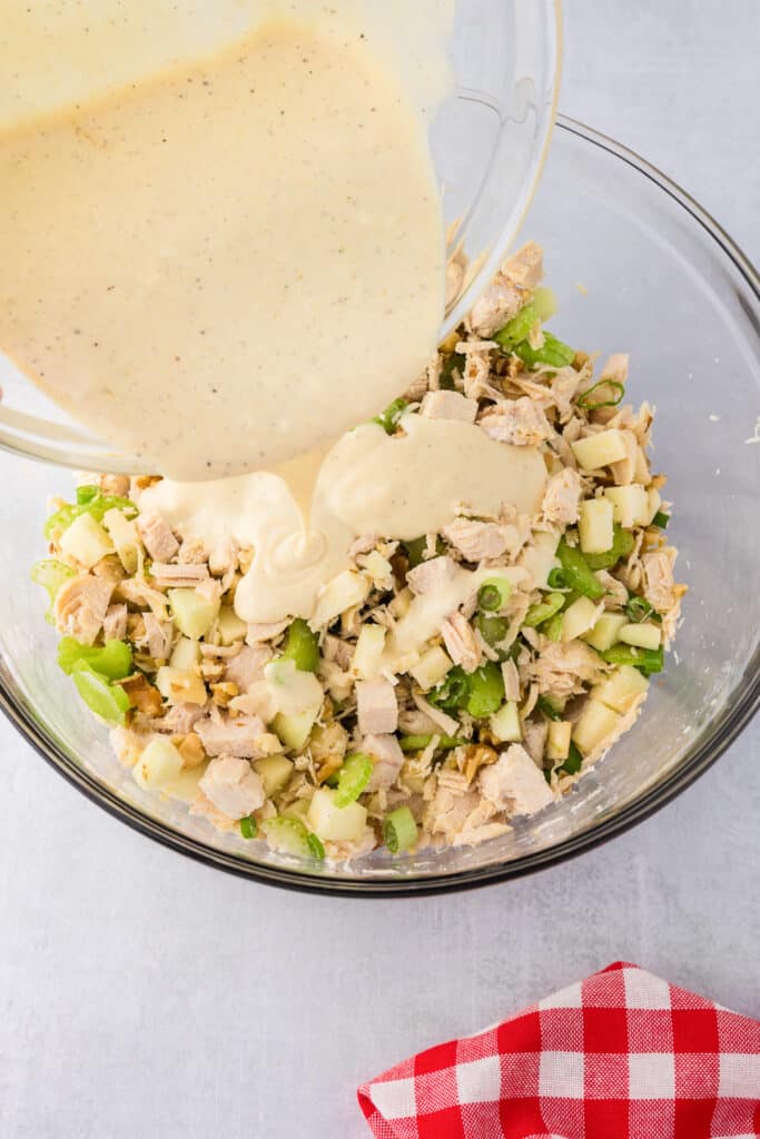 Dressing being poured into a bowl of chopped chicken, apples and celery pieces to make apple walnut chicken salad.