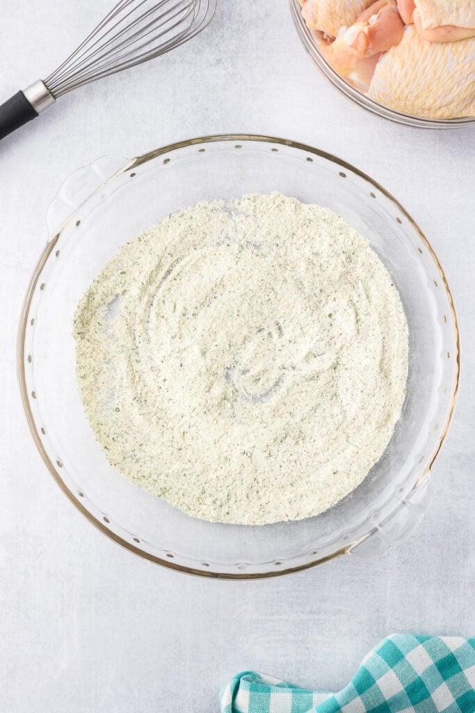 A glass bowl containing seasoning and baking powder for crispy ranch chicken thighs sits on a counter next to raw chicken thighs in a bowl.