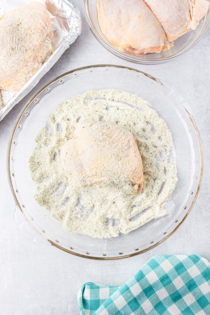 Raw bone in chicken thighs being coated in ranch seasoning and baking powder in a glass bowl and being placed on a baking sheet nearby.