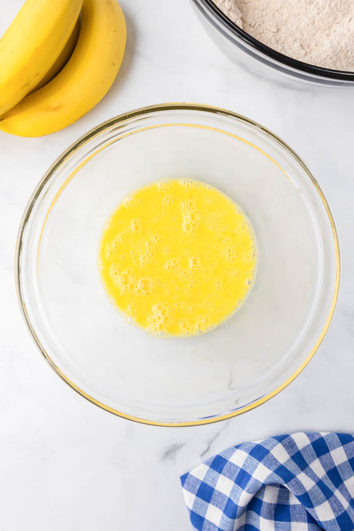 Whisked eggs in a clear glass bowl on a kitchen counter with bananas and ingredients in the background.