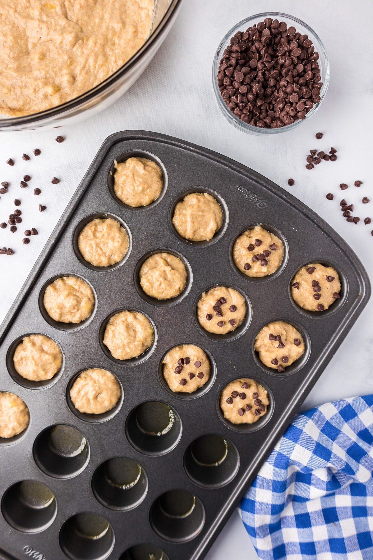 Unbaked muffin banana chocolate chip mini muffins batter being filled in a muffin tin with chocolate chips being sprinkled on top.