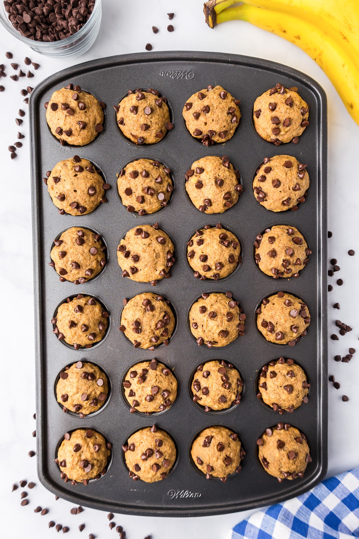 A tray of freshly baked Banana chocolate chip mini muffins with loose chocolate chips and bananas on the side of the pan.