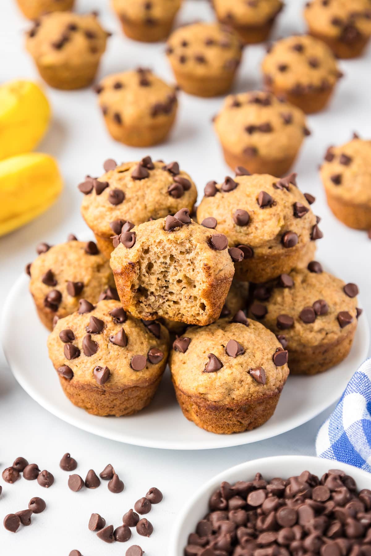 Plate of freshly baked mini banana chocolate chip muffins surrounded by additional muffins, chocolate chips, and bananas on a kitchen counter.