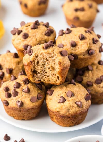 Banana chocolate chip mini muffins piled high on a plate with one muffin missing a bite.