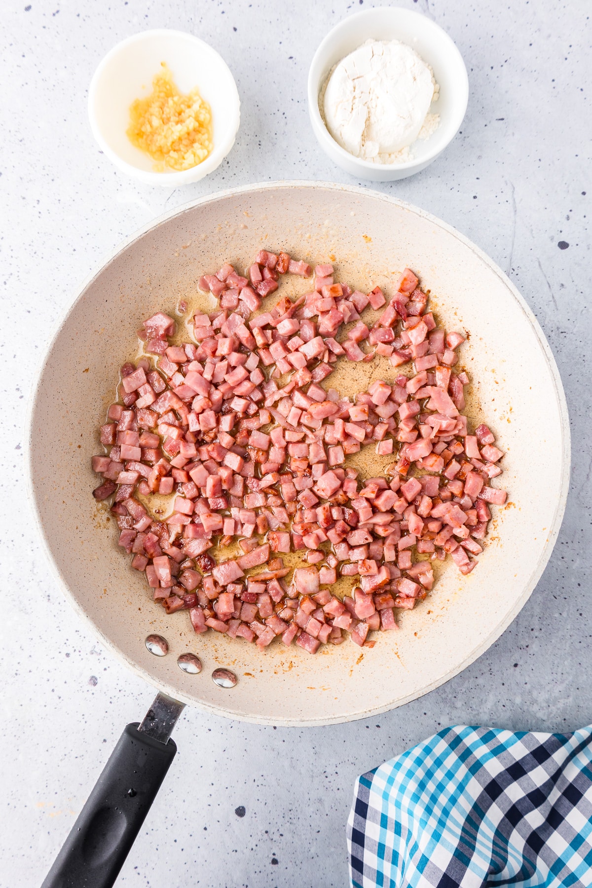 Diced ham being browned in a medium frying pan with flour and minced garlic nearby on the counter in bowls.