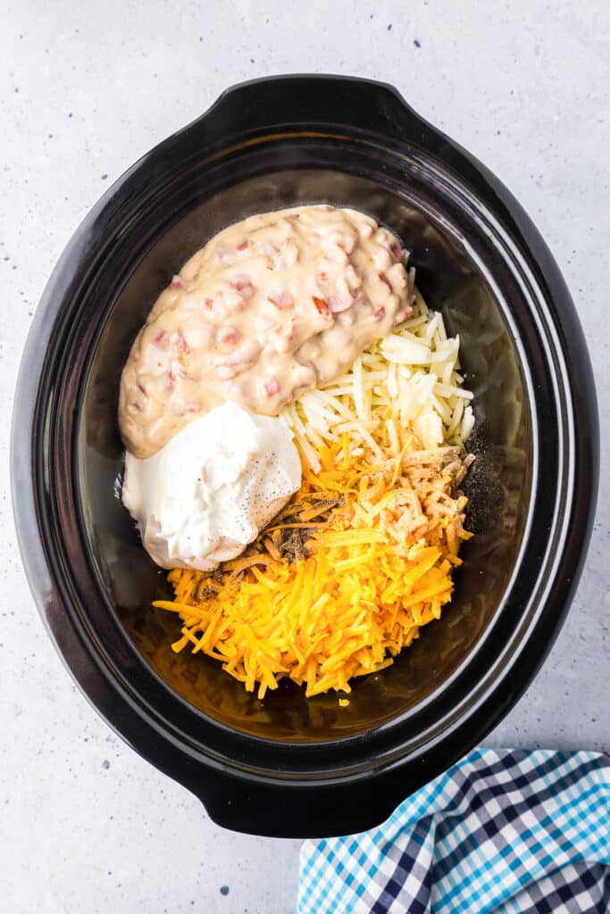 Hash brown casserole ingredients such as the ham and sauce mixture, sour cream, shredded potatoes and cheese in a slow cooker before mixing.