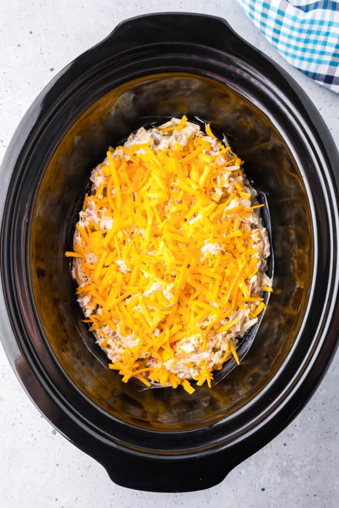 Hash brown casserole in a slow cooker base topped with shredded cheese before cooking.