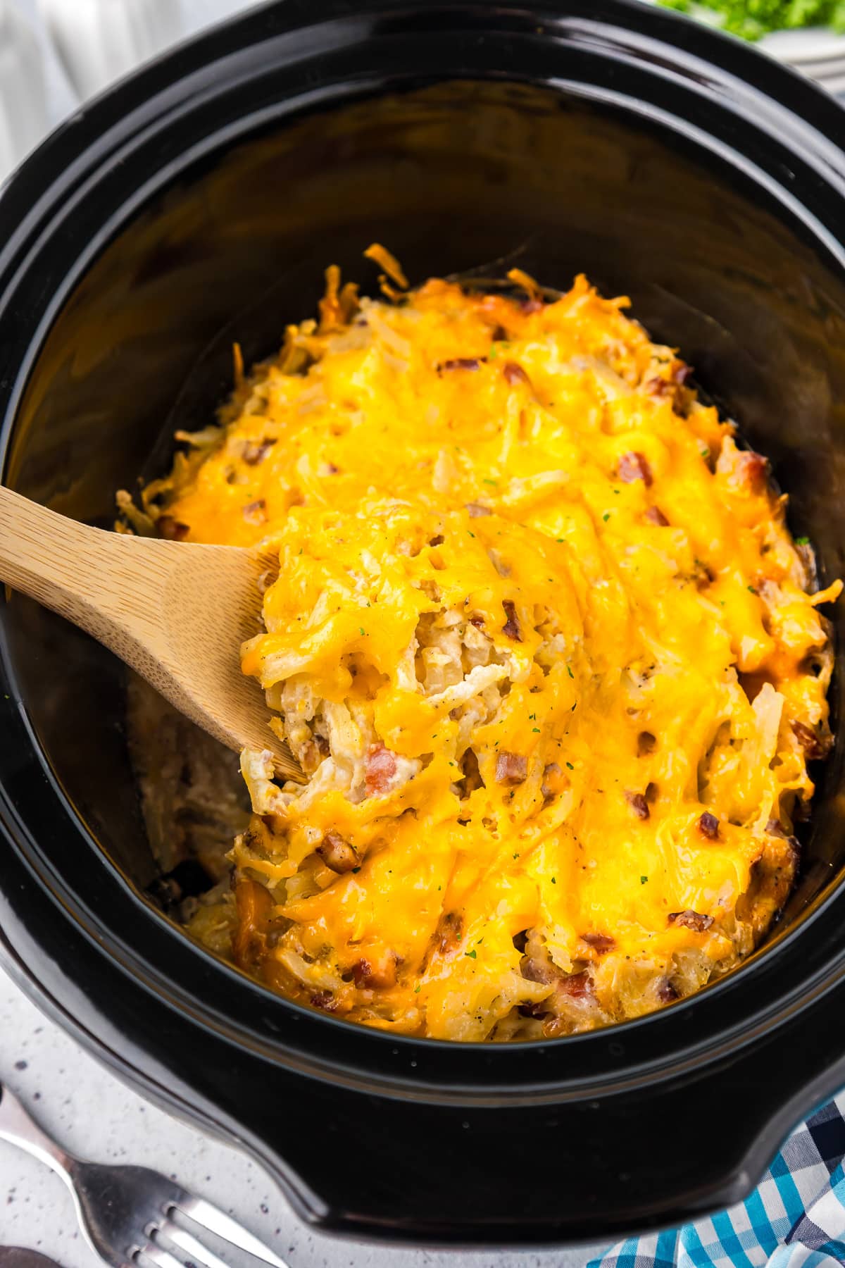 A slow cooker filled with a cheesy hashbrown casserole topped with melted cheddar cheese being scooped and a wooden spoon.