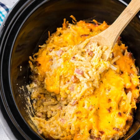 A slow cooker filled with cheesy hashbrown casserole with bits of ham and a wooden spoon with a big scoop.