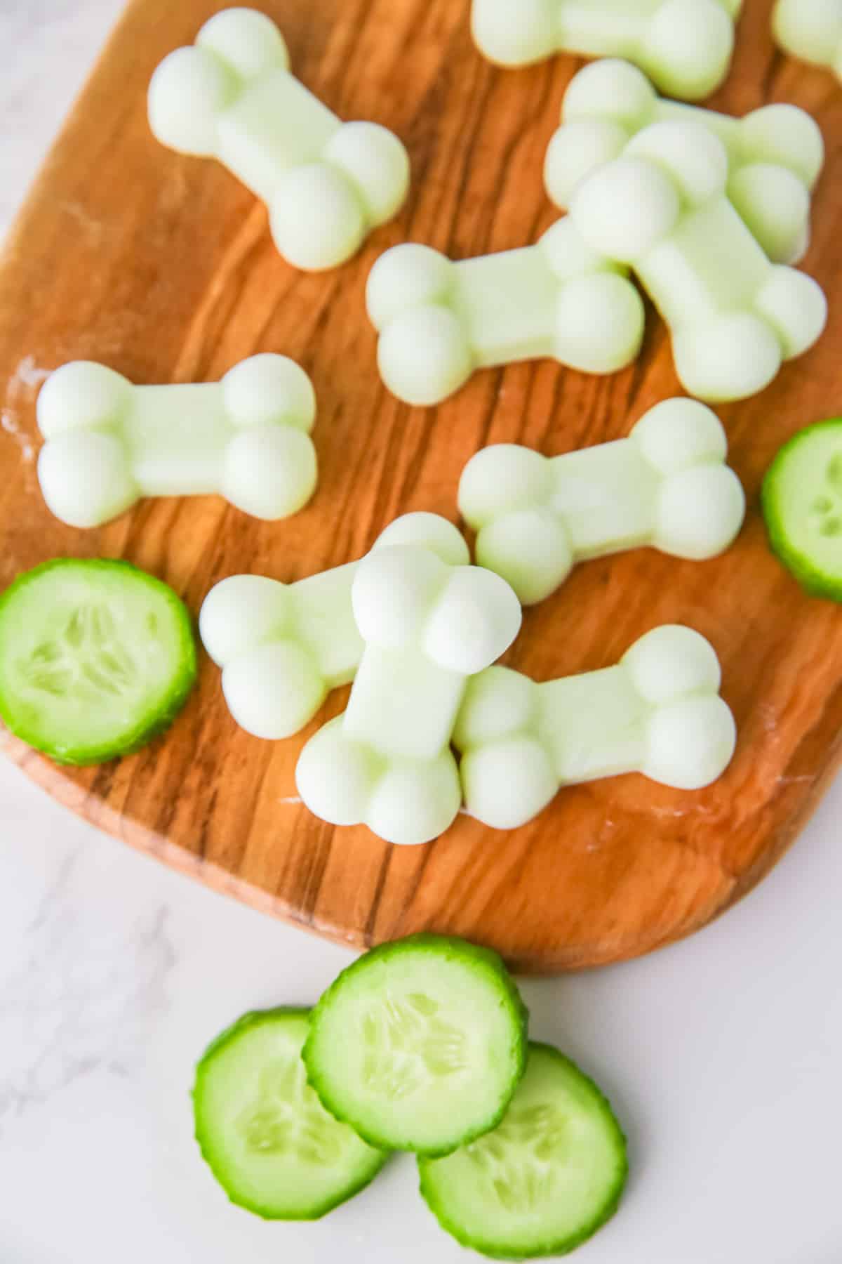 Bone-shaped yogurt cucumber dog treats and slices of cucumber on a wooden cutting board.