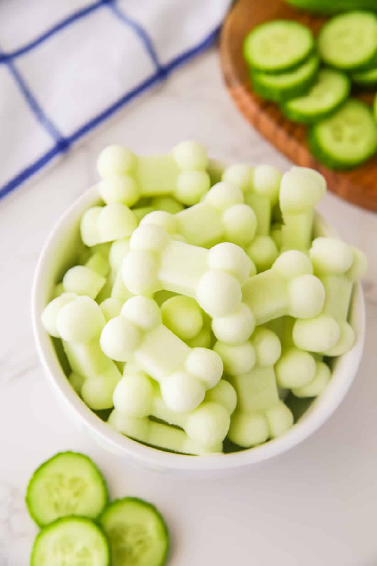 A bowl of bone-shaped yogurt cucumber treats on a white surface, with more cucumber slices and a blue-striped towel in the background.
