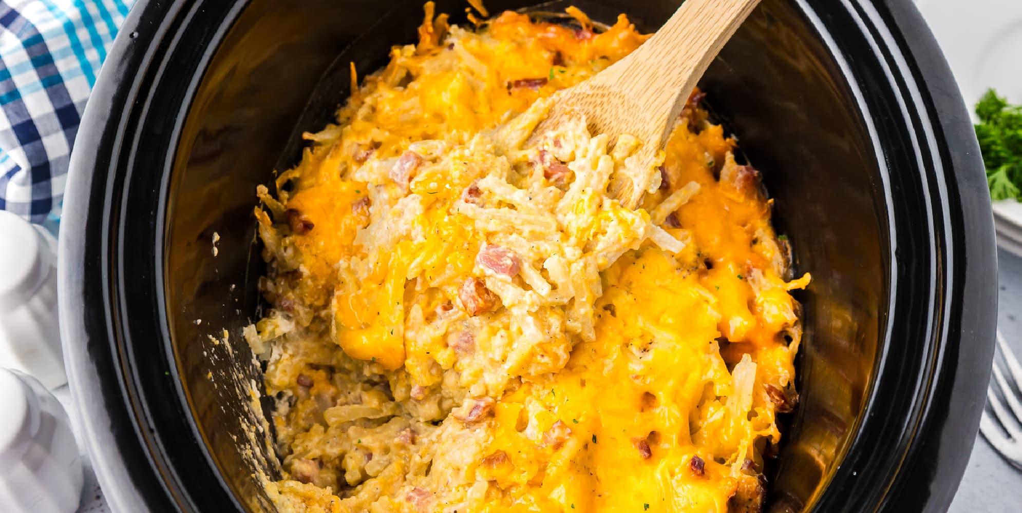 Wide view of cheesy hash brown casserole with pieces of ham in a slow cooker with a wooden spoon stirring the potatoes.