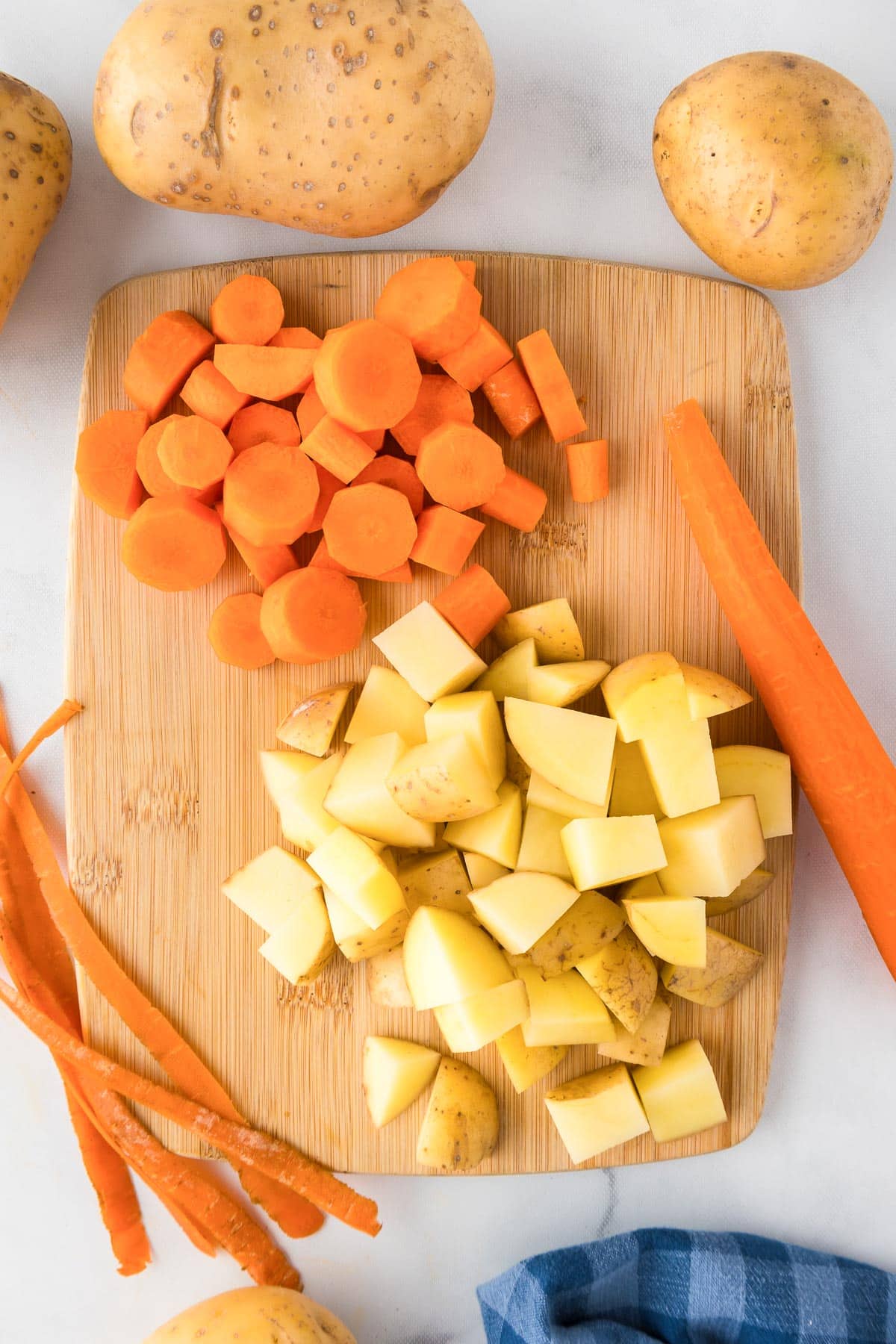Chopped potatoes and carrots into chunks on a wooden cutting board, with whole vegetables and a cloth nearby on the countertop.