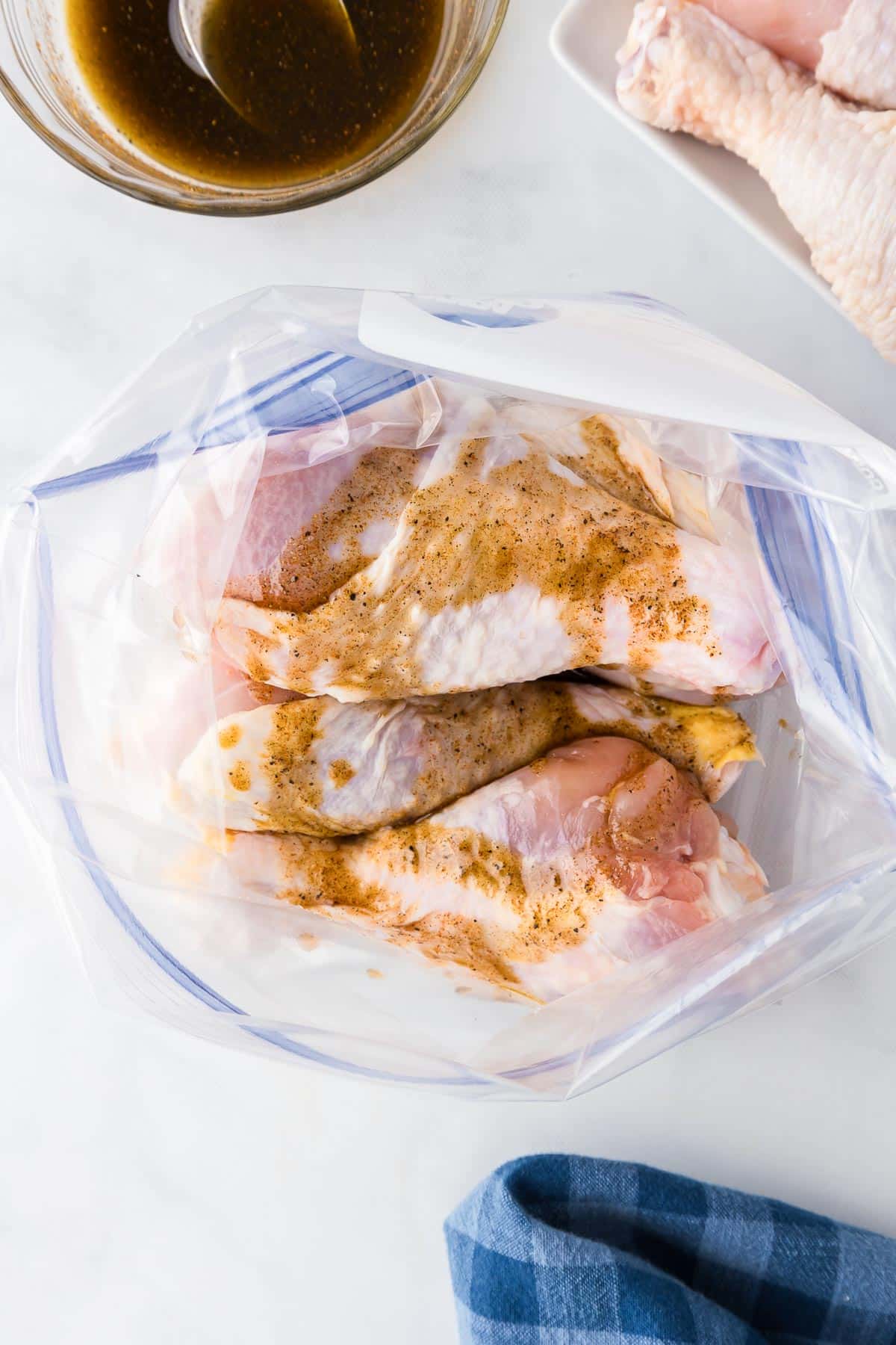 Raw chicken legs in a zip-top bag seasoned with some marinade on a kitchen counter, with a bowl of marinade nearby.