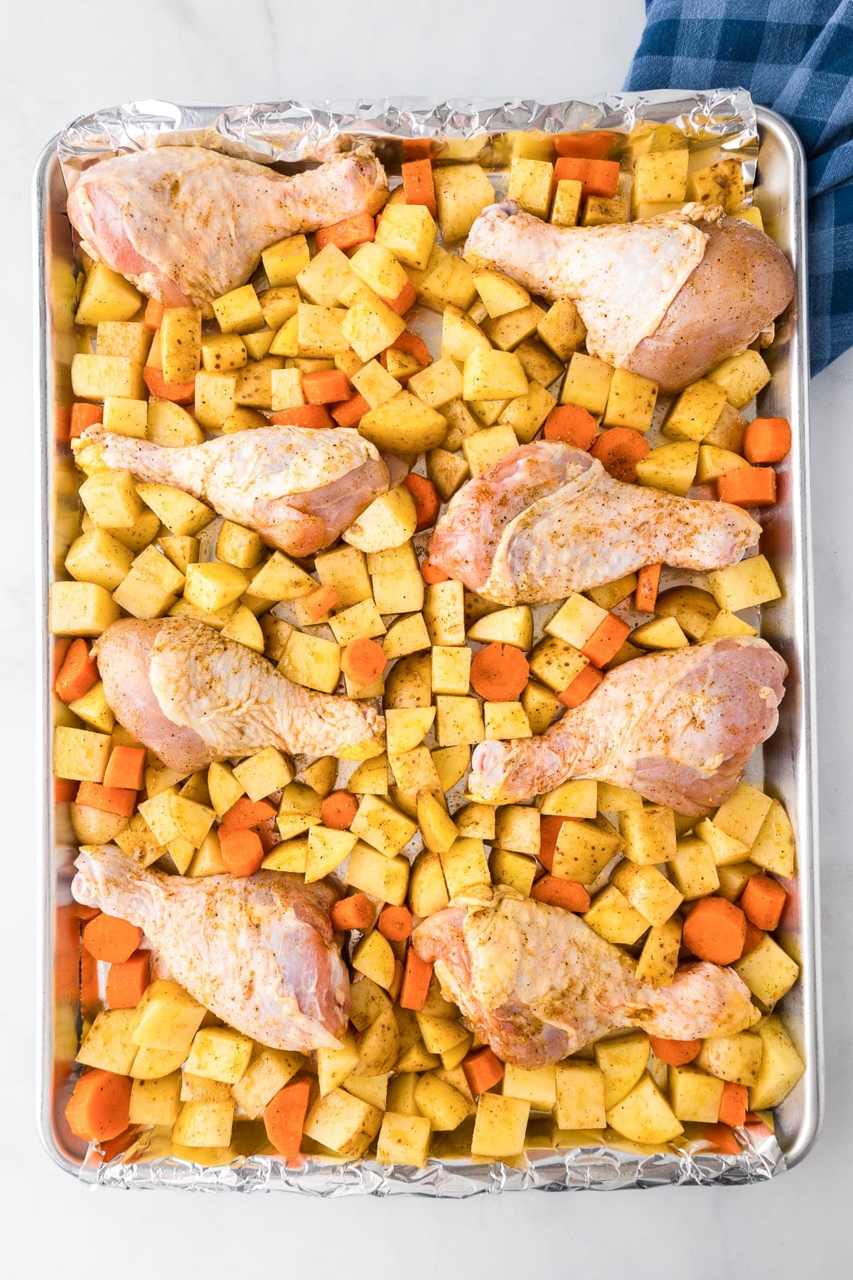 Raw chicken legs and diced vegetables seasoned and arranged in a baking tray, ready for cooking.