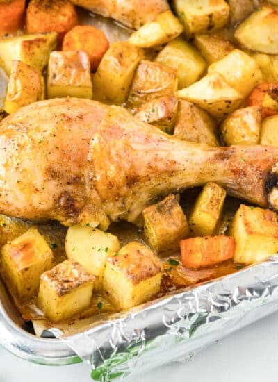 Roasted chicken leg on a bed of golden potatoes and carrots on a foil-lined baking tray from the side.