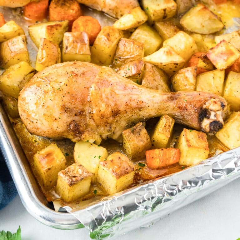 Roasted chicken leg on a bed of golden potatoes and carrots on a foil-lined baking tray from the side.