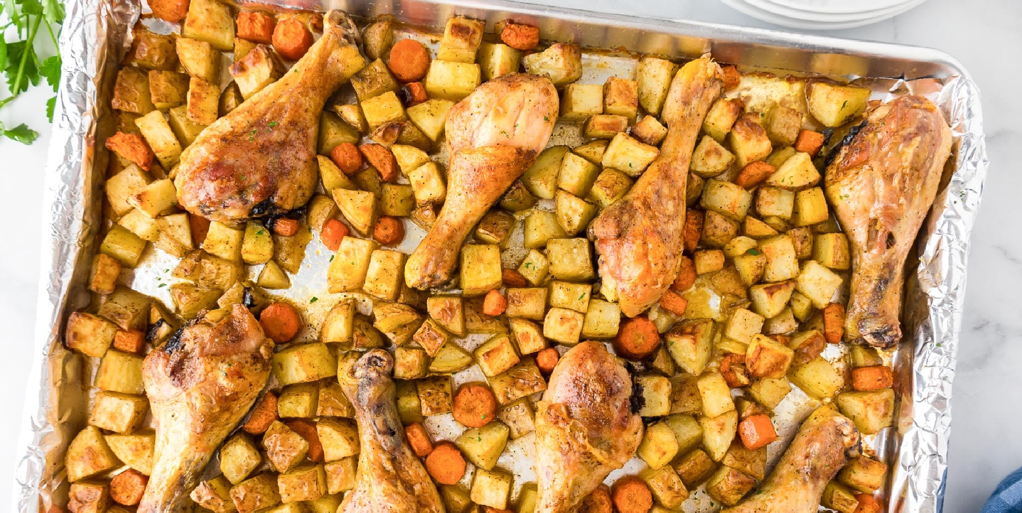 Oven-roasted chicken legs and mixed vegetables including diced potatoes and sliced carrots on a foil-lined baking tray.