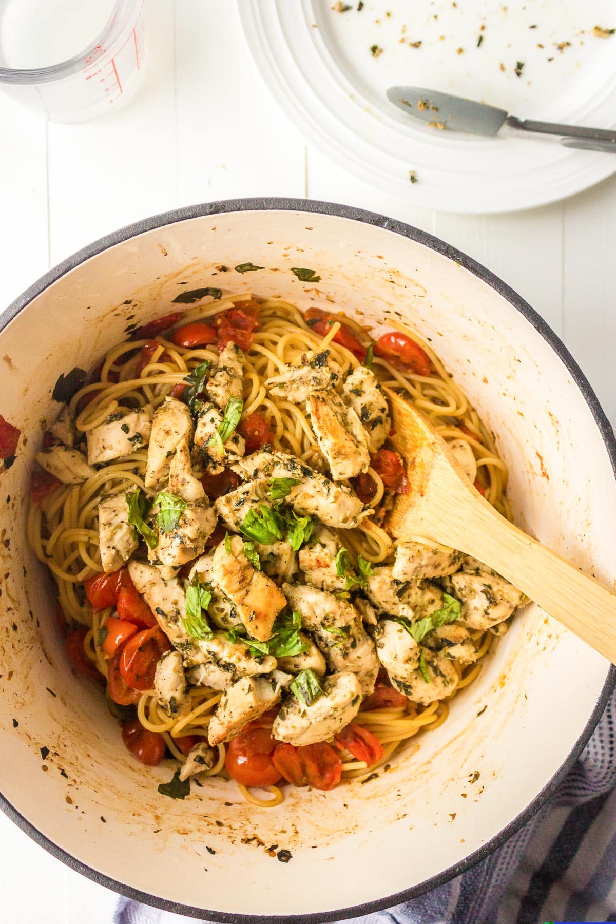 A pot of spaghetti with grilled chicken, cherry tomatoes, and basil, served with a wooden spoon, on a white tabletop.