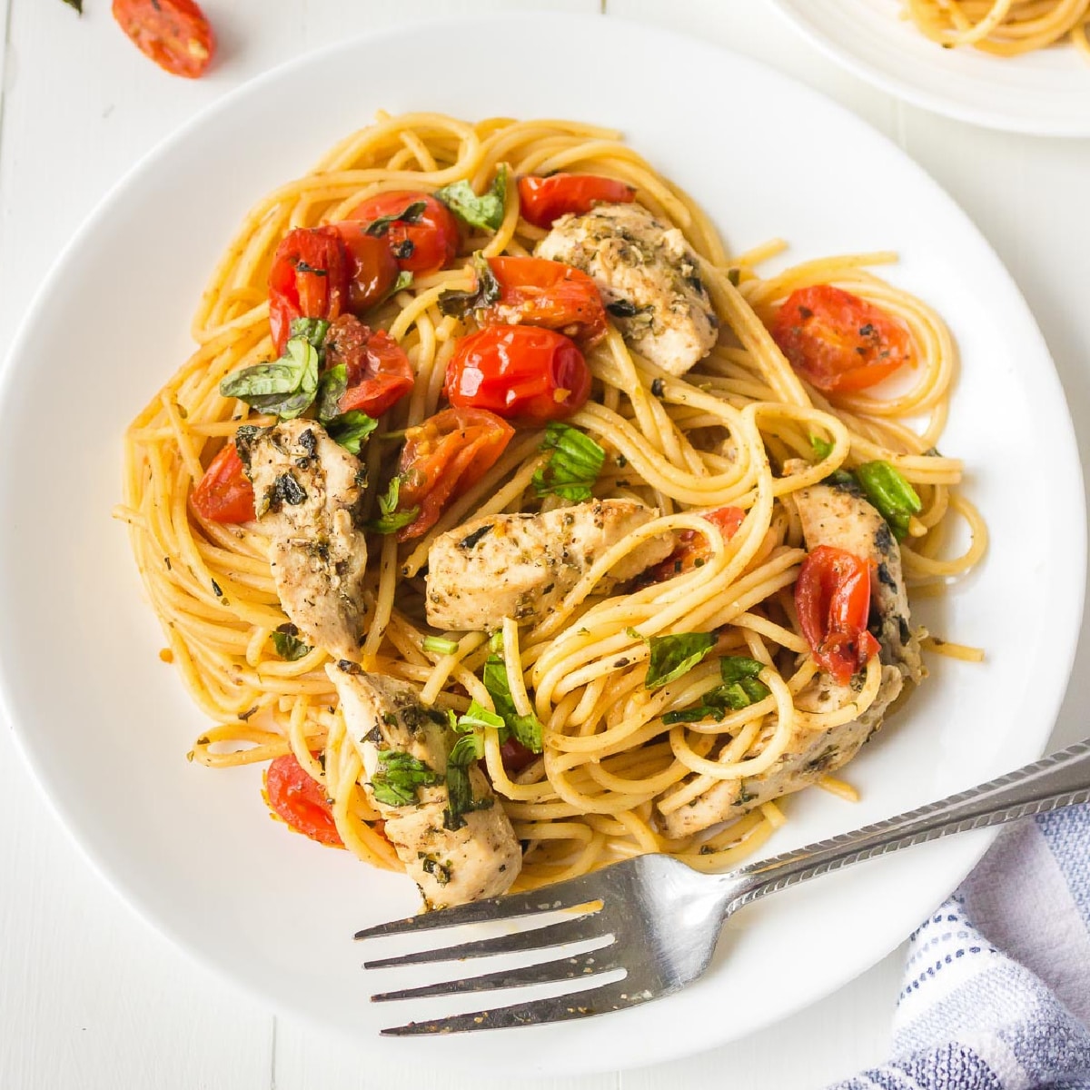 Close-up of a plate of bruschetta chicken pasta spaghetti with herbed chicken pieces, cherry tomatoes, and fresh basil on a plate with a fork.