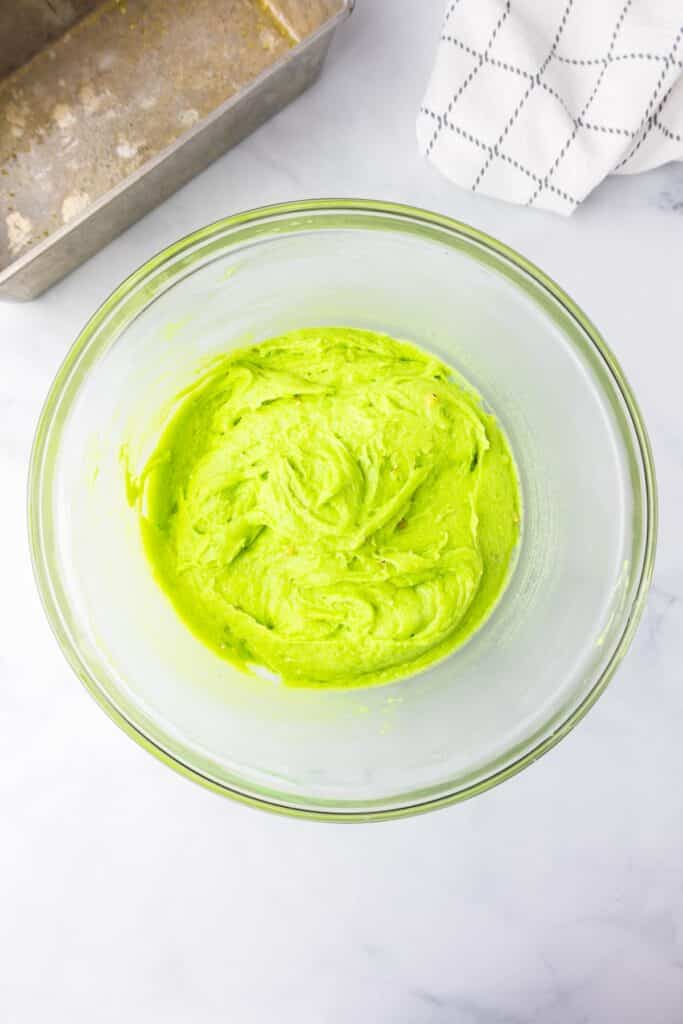 A bowl of vibrant green pistachio bread batter on a kitchen counter next to a bread pan and a white cloth.