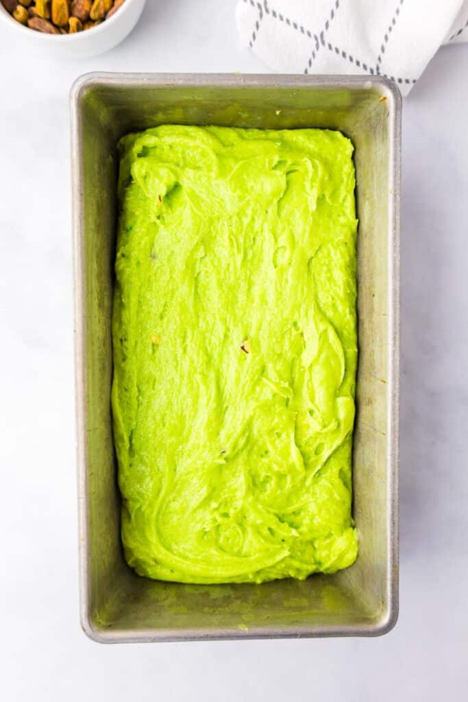 Unbaked pistachio bread dough in a metal loaf pan on a white surface, with pistachio nuts in a bowl, and a cloth.