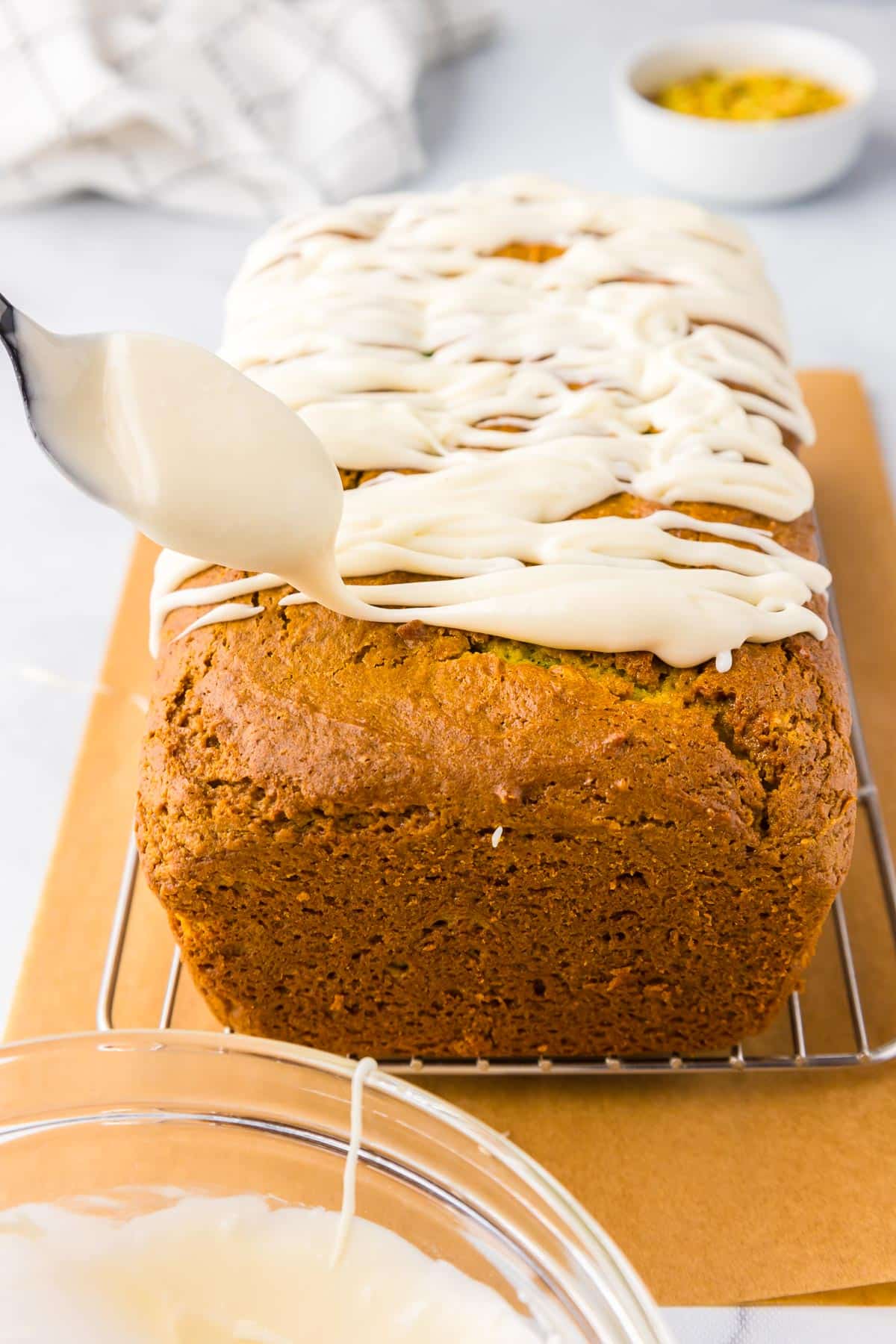 A loaf of freshly baked pirtachio bread with white icing being drizzled on top over a wire cooling rack.