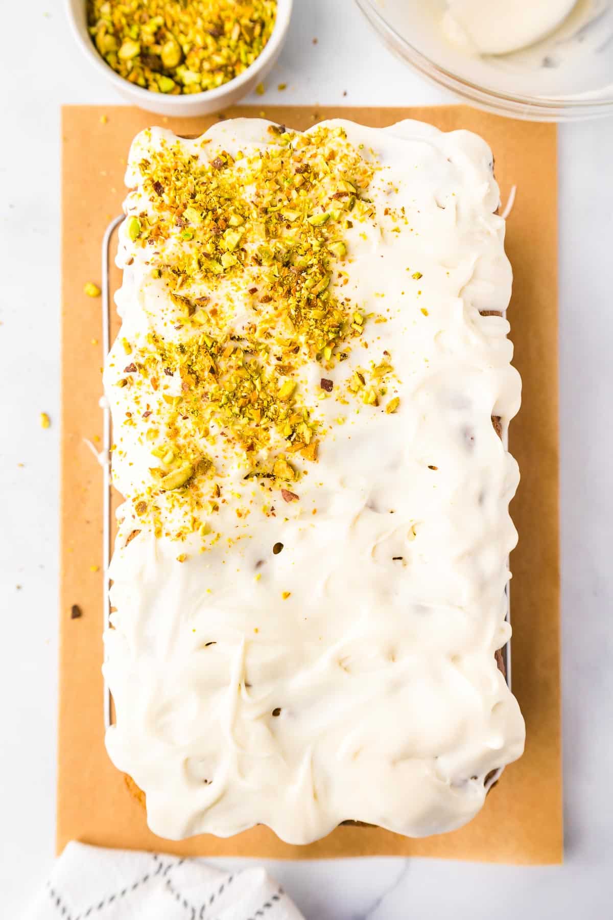 A pistachio loaf cake topped with white icing being sprinkled with crushed pistachios on a cutting board with a bowl of pistachios nearby.