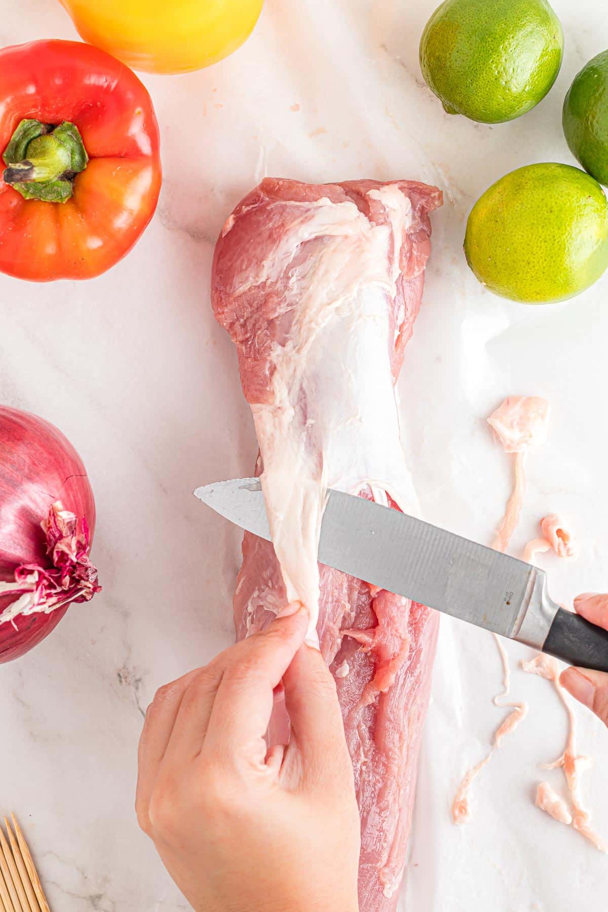 A person trimming the silver skin from a raw pork tenderloin with a knife on a counter.