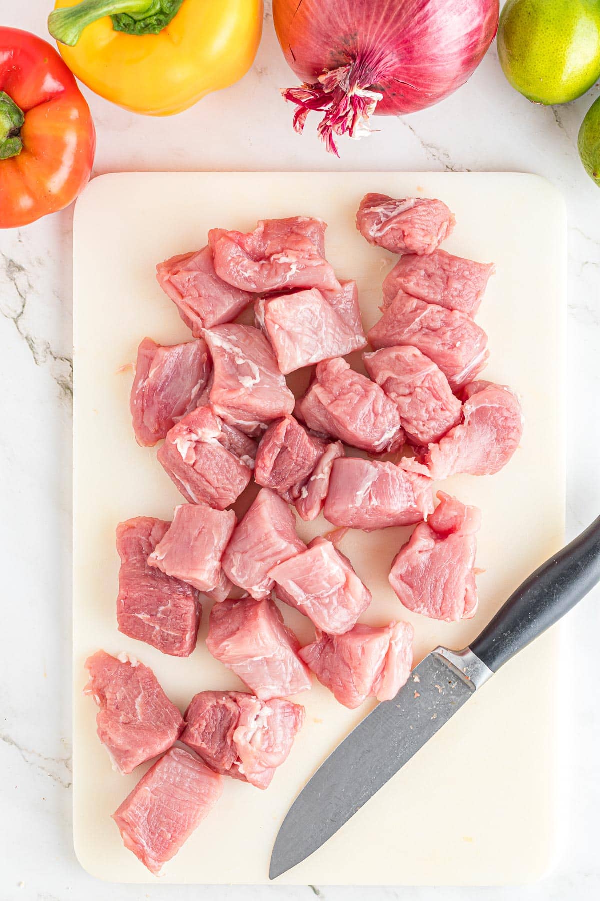 Raw pork tenderloin pieces on a white cutting board with a knife and assorted vegetables in the background.