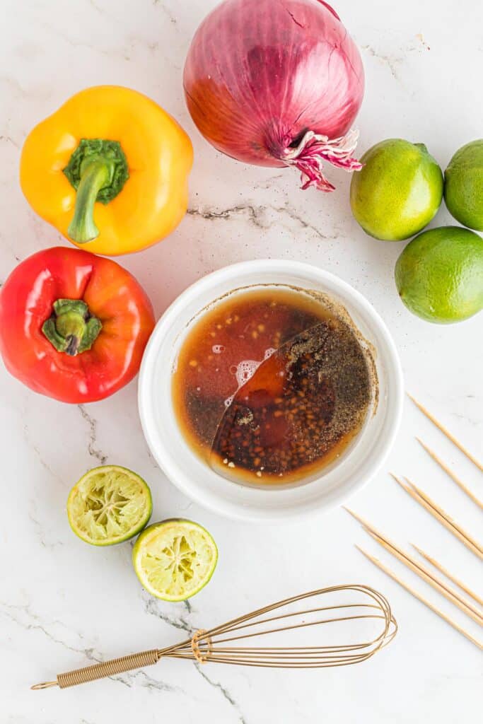 Top-down view of a bowl of soy sauce and other marinade ingredients next to a whisk surrounded by bell peppers, onions, limes, and skewers.