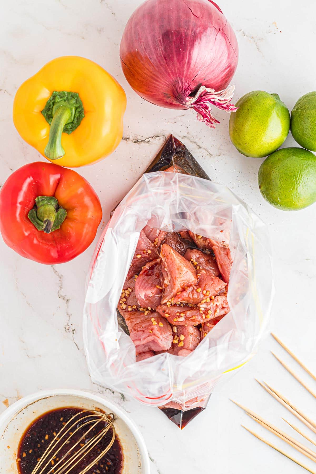 Overhead view of pork tenderloin pieces in a bag full of marinade with more marinade and skewer vegetables nearby.