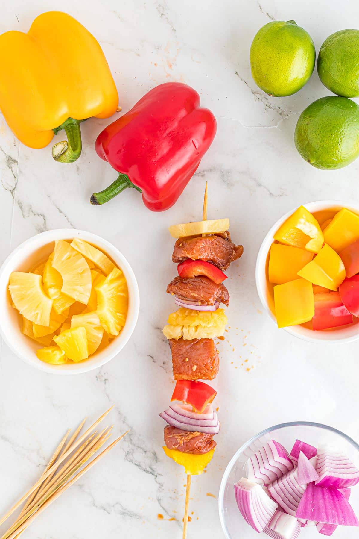 Ingredients for making skewers in bowls, including chopped bell peppers, onions, pineapple and an assembled skewer that has pieces of raw pork tenderloin and vegetables on it.