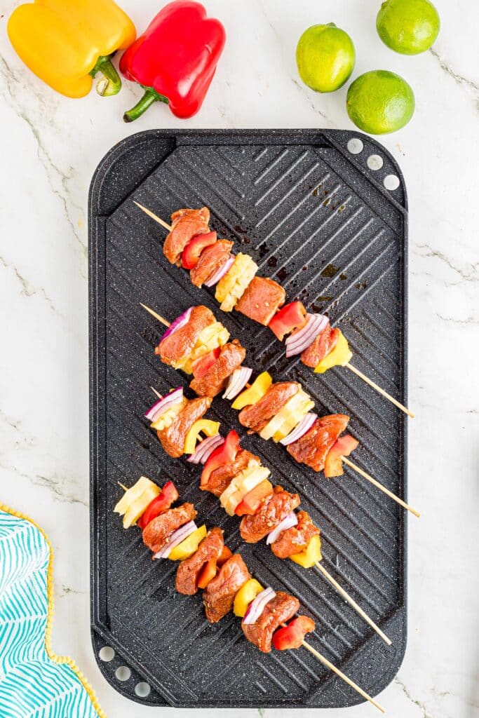 Pork tenderloin and vegetable skewers with chunks of pork, bell peppers, and pineapple on a black grill pan as it starts to grill.