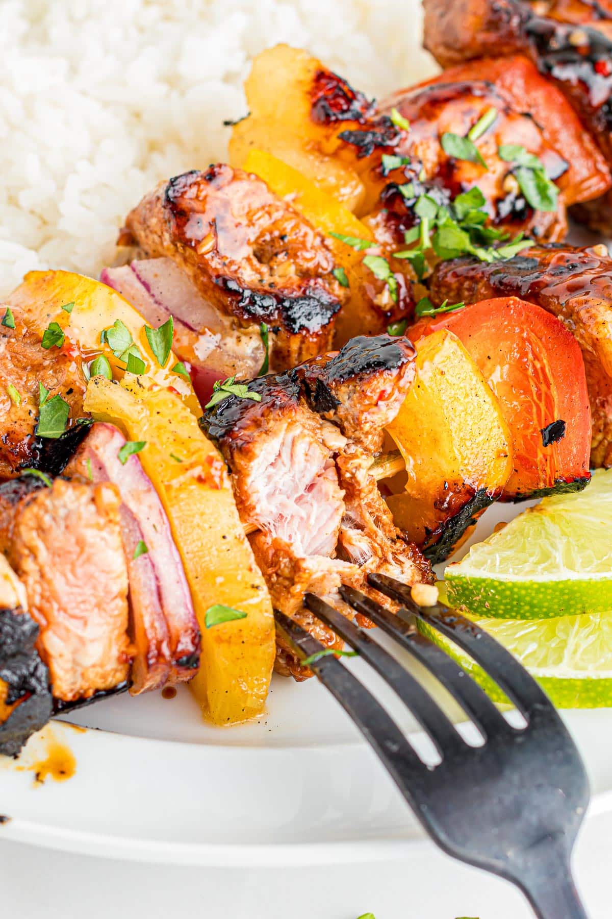 Grilled pork skewers with pineapple and bell peppers with a fork showing the inside of a piece of pork on a plate next to rice.