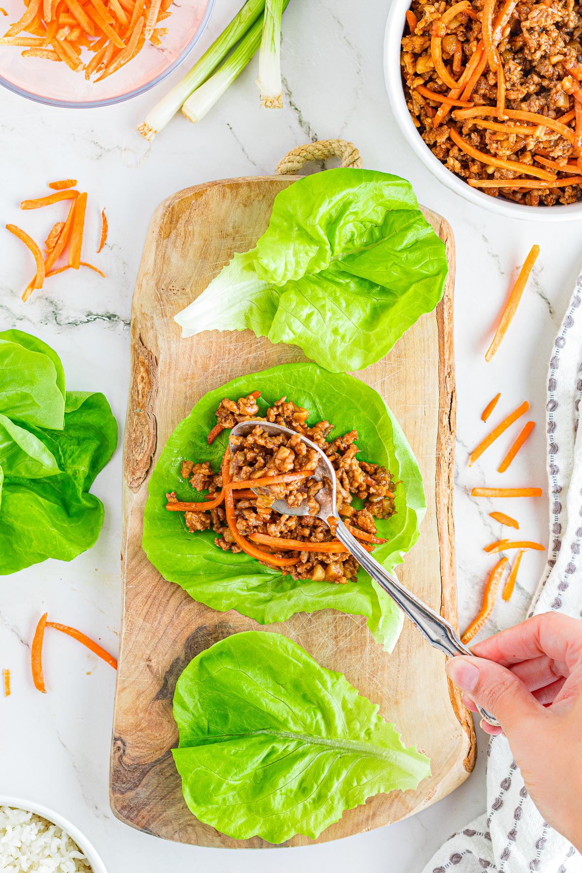 A hand filling asian-style pork lettuce wraps wwith ground pork and vegetables being spooned into lettuce pieces on a wooden cutting board.