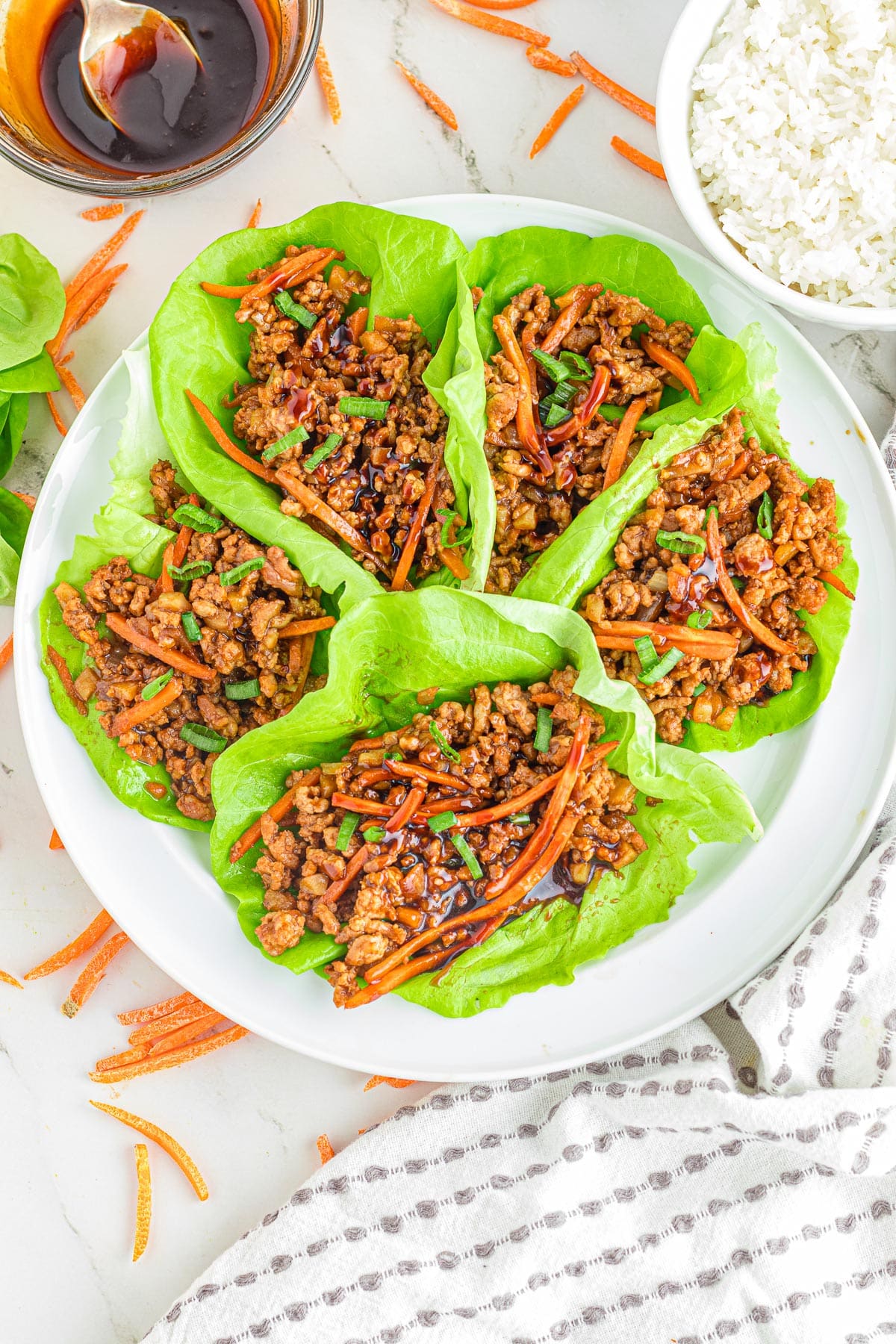 Pork lettuce wraps filled with seasoned ground meat and carrots being served on a platter with more sauce and white rice in a bowl nearby.