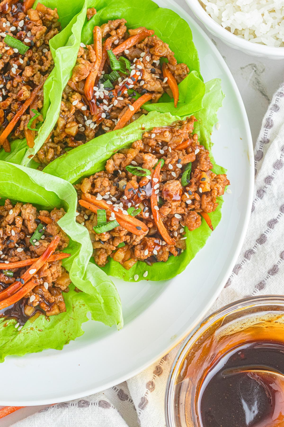 Asian-style pork lettuce wraps filled with ground meat and vegetables, garnished with sesame seeds, served with rice and dipping sauce on the side of a plate.