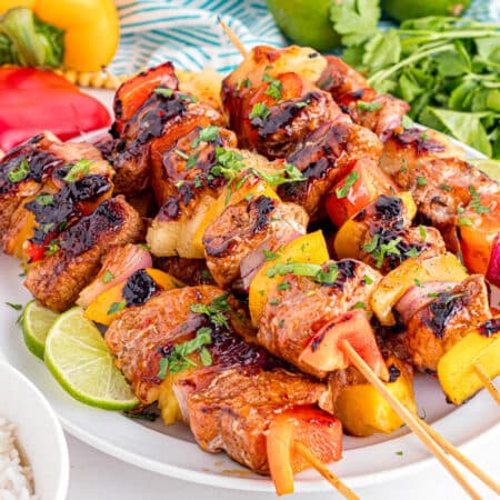A plate of grilled pork tenderloin skewers with chunks of pork, bell peppers, pineapple, and onions, garnished with herbs and lime wedges.