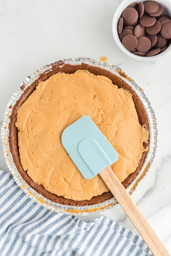 A pie with an orange filling is in a foil pie crust. A light blue spatula is resting on top. A bowl of chocolate wafers and a striped cloth are nearby.