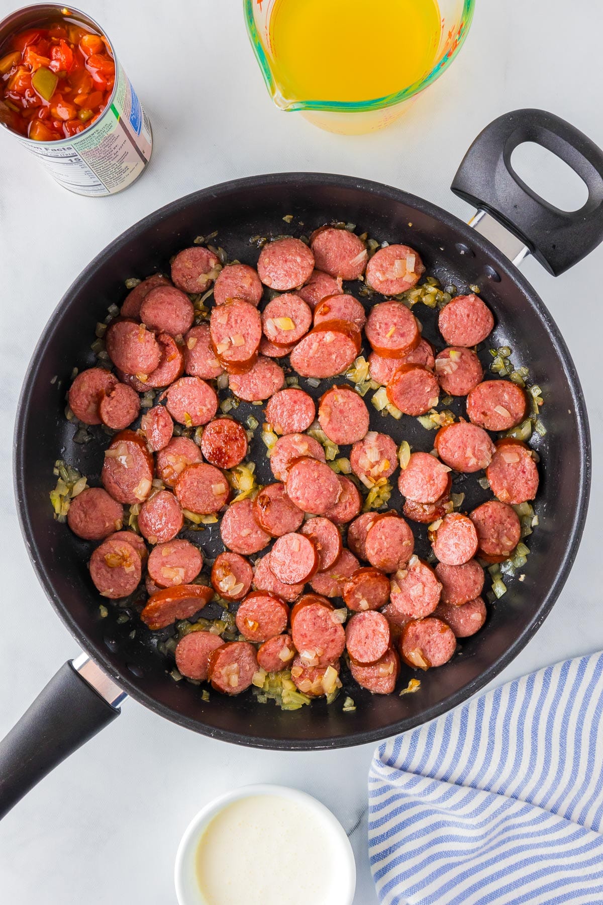 A frying pan contains browned sliced sausages and onions. Nearby on the counter are a can of green chili diced tomatoes, chicken broth and a bowl of heavy cream.