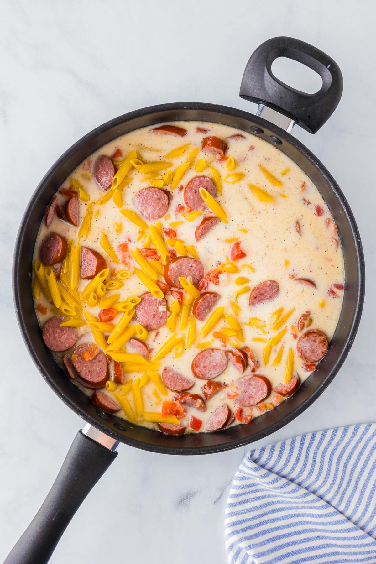 A pan on a stovetop contains a creamy sauce mixed with sliced sausages, pasta, and bits of vegetables. A striped cloth is partially visible in the bottom right corner.