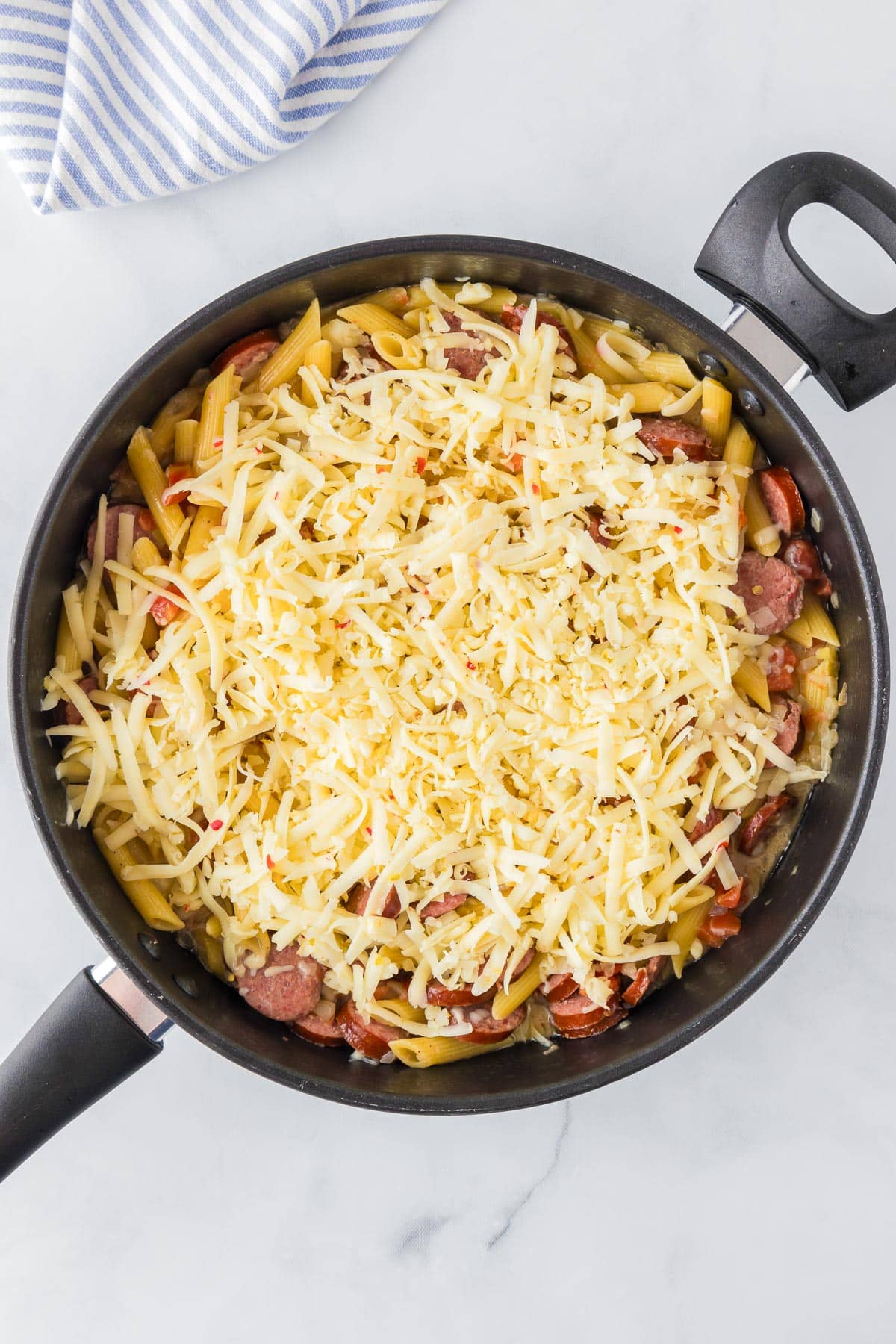 A skillet filled with penne pasta, sausage slices, and shredded cheesy to make cheesy smoked sausage pasta.