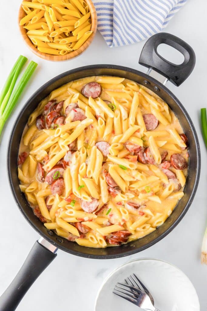 A skillet with creamy cheesy smoked sausage pasta with a bowl with forks, green onion and pasta in a bowl nearby.
