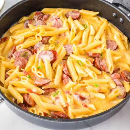 A pan filled with cooked penne pasta mixed with sliced sausages and topped with a creamy cheese sauce to make a one pot sausage pasta.