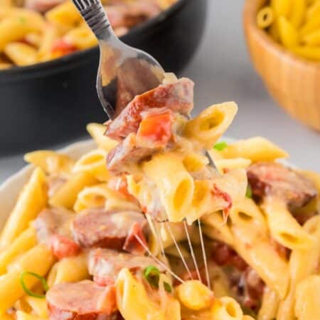 A fork holds a bite of creamy cheesy smoked sausage pasta with more pasta in the pan in the background.