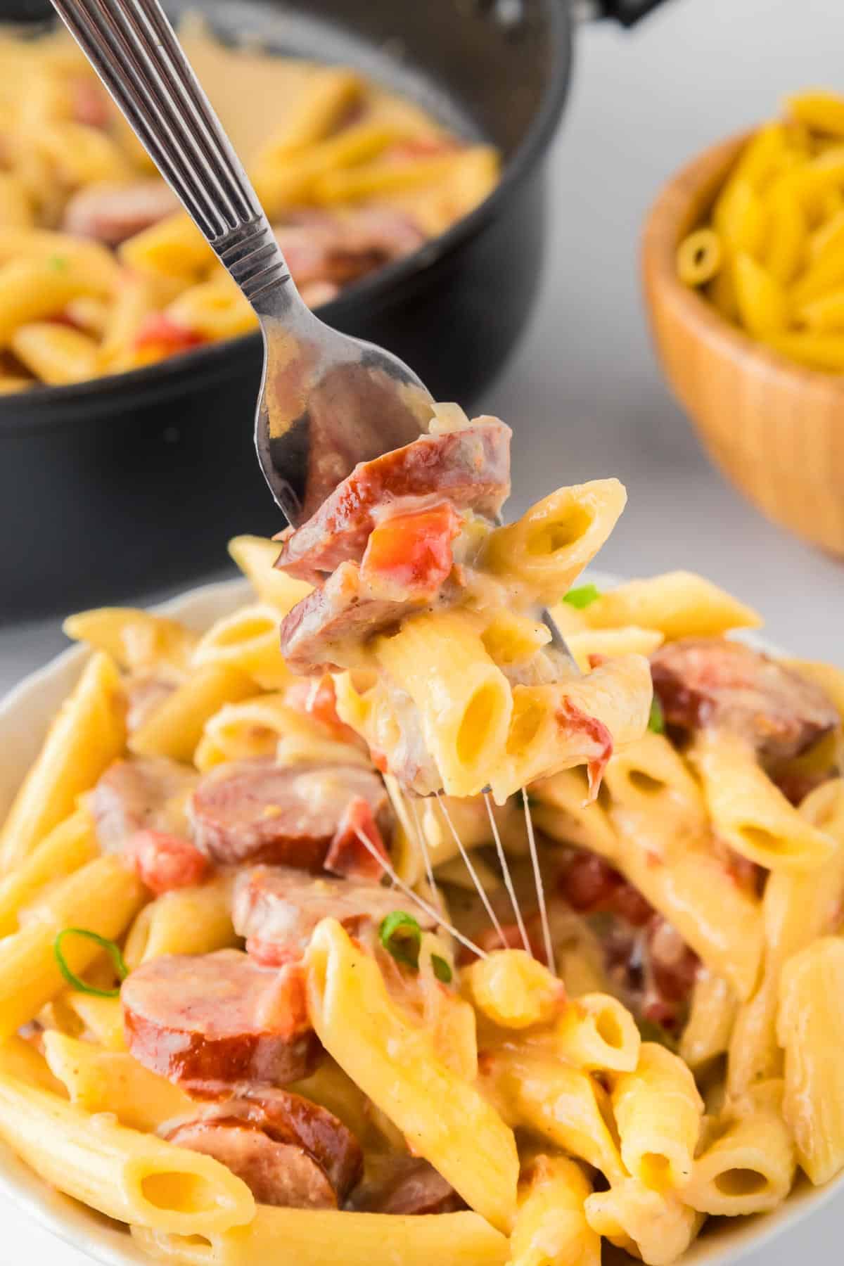 A fork holds a bite of creamy cheesy smoked sausage pasta with more pasta in the pan in the background.