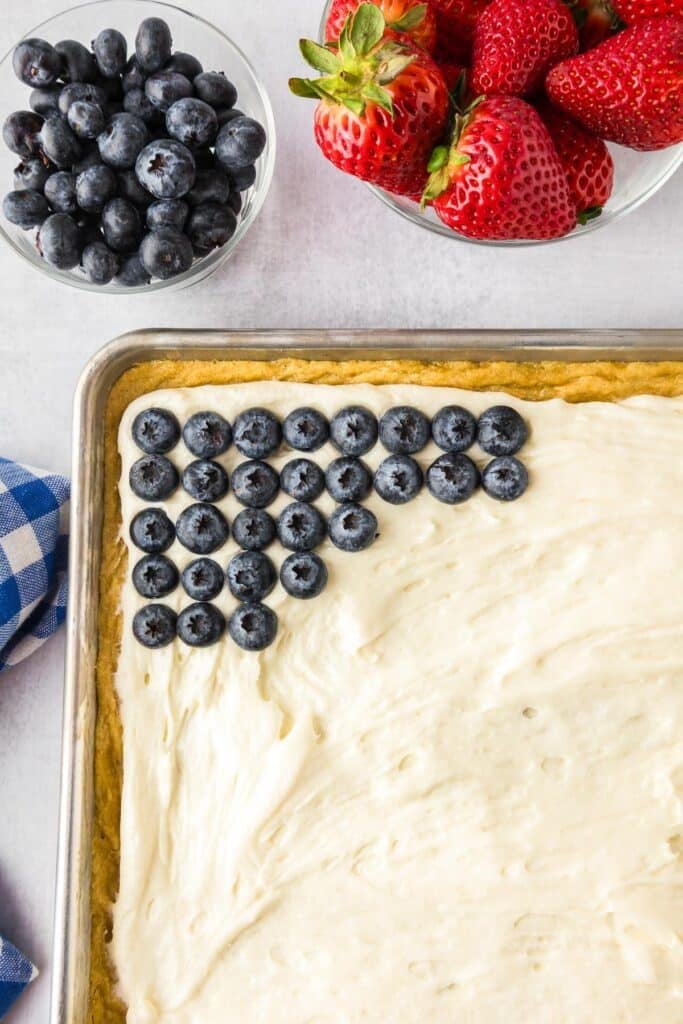 A rectangular baking pan with cream cheese frosting focused on the corner using blueberries to make the blue rectangle of the flag fruit pizza.